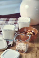 Three glass jars of homemade yogurt and a wooden bowl with nuts on a wooden board with two caps. White jug of milk.