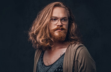 Fototapeta premium Close-up portrait of a redhead hipster male with long luxuriant hair and full beard dressed in casual clothes standing in a studio, looking away. Isolated on the dark background.