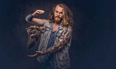 Portrait of a tattoed redhead hipster male with long luxuriant hair and full beard dressed in a t-shirt and jacket holds a keeps the scarecrow of an owl in a studio. Isolated on the dark background.