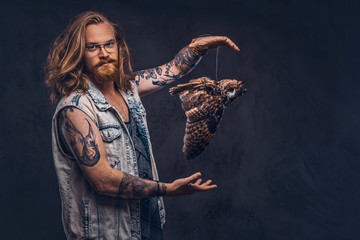 Portrait of a tattoed redhead hipster male with long luxuriant hair and full beard dressed in a t-shirt and jacket holds a keeps the scarecrow of an owl in a studio. Isolated on the dark background.