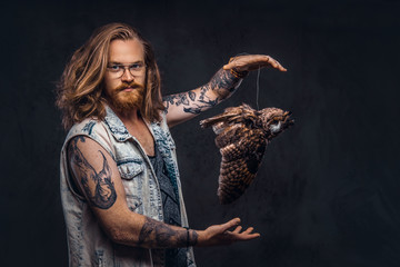 Portrait of a tattoed redhead hipster male with long luxuriant hair and full beard dressed in a t-shirt and jacket holds a keeps the scarecrow of an owl in a studio. Isolated on the dark background.