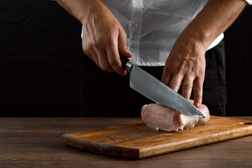 The chef cuts a piece of fresh meat on a wooden board against a dark background, hands close-up. The concept of cooking, cooking meat, a recipe for meat dishes.