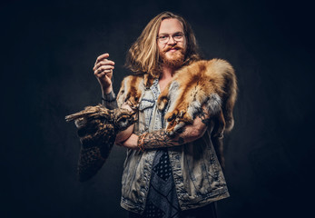 Portrait of tattoed redhead hipster male with long luxuriant hair and full beard dressed in a t-shirt and jacket holds a keeps the scarecrow of an owl and fox skin on a shoulder in a studio.