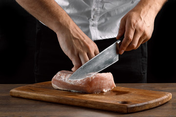 The chef cuts a piece of fresh meat on a wooden board against a dark background, hands close-up. The concept of cooking, cooking meat, a recipe for meat dishes.
