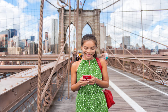 NYC travel phone texting girl holding smartphone on Brooklyn bridge in New York City, Manhattan USA. Asian woman phone reading or using social media in summer.