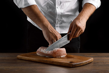 The chef cuts a piece of fresh meat on a wooden board against a dark background, hands close-up. The concept of cooking, cooking meat, a recipe for meat dishes.