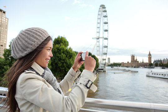 Tourist Asian girl taking picture with phone of London city landscape with popular attraction at Thames River. Europe travel lifestyle. Happy woman holding cellphone outdoors in spring coat.