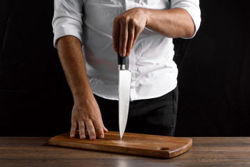 Hands of the chef close-up with a knife and a wooden board on a dark background. The concept of cooking, cooking, recipe dishes.