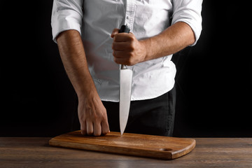 Hands of the chef close-up with a knife and a wooden board on a dark background. The concept of cooking, cooking, recipe dishes.