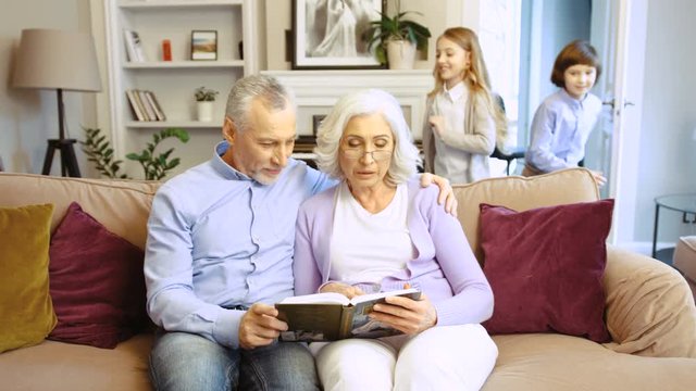 Grandparents reading boook on the sofa at home in the living room while their grandson and granddaughter running to the room at sit near them on the sofa.