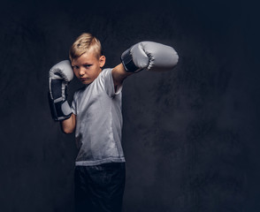 Schoolboy boxer with blonde hair dressed in a white t-shirt wearing boxing gloves shows a boxing kick. Isolated on a dark textured background.