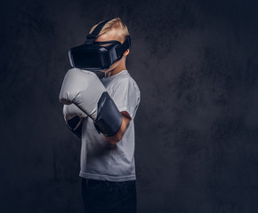 Schoolboy boxer with blonde hair dressed in a white t-shirt wearing visual reality glasses and boxing gloves, workout in a studio. Isolated on dark textured background.