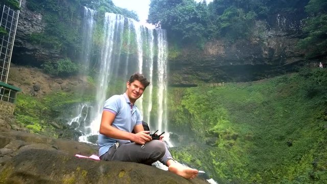 guy sits on rock with flycam remote control smiling by waterfall
