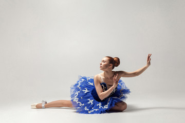 Naklejka premium attractive ballerina with bun collected hair wearing blue dress and pointe shoes graceful sitting on white studio floor.