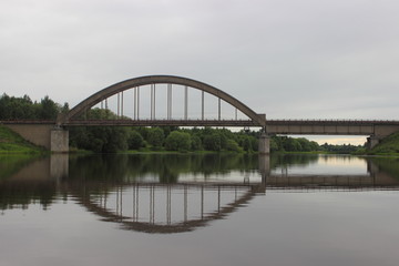 Fototapeta na wymiar Classic old arched railway bridge over the river - view from the water on a summer day against the trees on the shore and gray cloudy sky