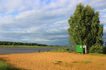 Deserted beach with dressing room and green birch in the wind in early autumn against the blue cloudy sky with gray clouds