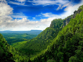 It is view of Pravcicka gate in national park in Hrensko in Czech republic. There is green forests and blue sky.  