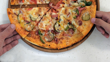 Close-up of pizza on a wooden plate. Two hands take pieces of pie with sausage, vegetables, cheese, spices in the dough.