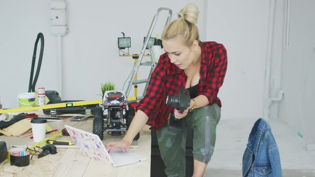 Beautiful young blond woman in bright casual clothes standing comfortably at workbench with tools and instruments and using remote controller of radio-controlled toy car placed on desk nearby.