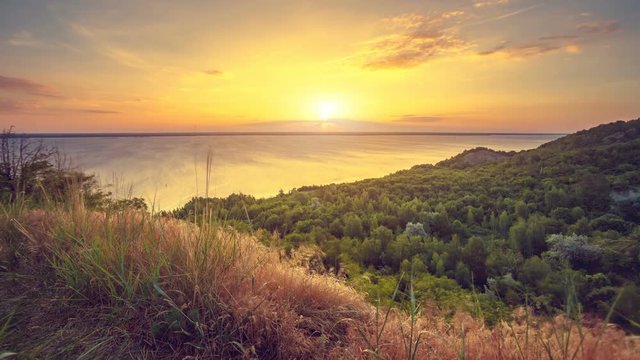 Time lapse sunset over beautiful seascape and mountain forest. Grass motion in the foreground. Autumn nature landscape. Bright warm toning filter. Holidays, travel, recreation. Ukraine, Europe. 4K