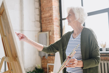 Waist up side view portrait of white haired senior woman holding palette painting pictures at easel...