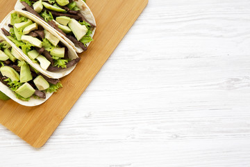 Corn tortillas with grilled beef, avocado and lime on the bamboo board over white wooden surface. View from above, overhead, flat-lay. Copy space and text area.