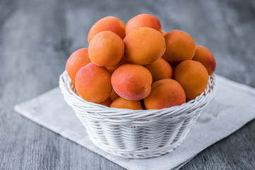 Ripe whole apricots in white basket on gray wooden background.