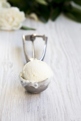 Vanilla ice cream in ice cream scoop with white peony flowers over white wooden surface, side view.
