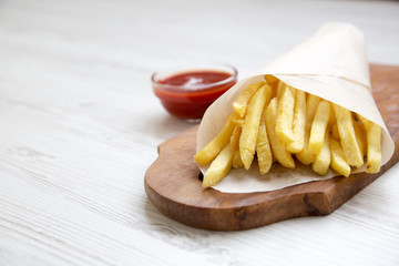 Fresh fried french fries with ketchup on wooden board over white wooden background, side view. Close-up. Copy space.