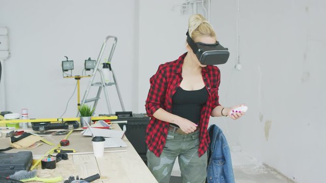 Cute blond young woman in casual clothes standing in virtual reality goggles headset at wooden workbench with tools and holding small white controller in hand 