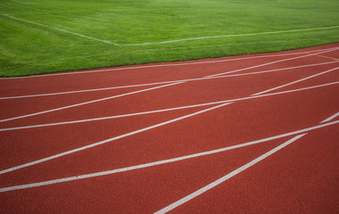 Fototapeta premium red running road track with white marking lines near green football field and empty space for copy or text