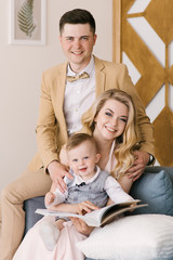 Obraz premium Beautiful young parents smile with their one-year-old child preview album at home in a beautiful interior in pastel colors. Family look.