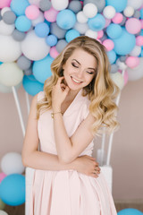 Obraz na płótnie Canvas Portrait of a beautiful young girl with curly blonde hair. Stands in a light dress against the background of white and blue balloons. Eyes look down with beautiful makeup closeup