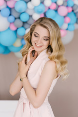 Portrait of a beautiful young girl with curly blonde hair. Stands in a light dress against the background of white and blue balloons. Eyes look down with beautiful makeup closeup