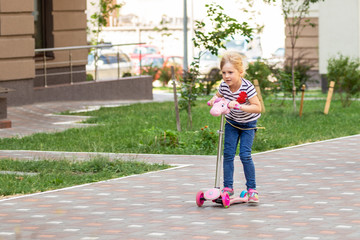 Cute little girl riding scooter in city park on bright summer day. Blond toddler having fun on street. Active leisure and outdoor sport for children concept