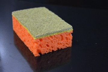 On a black glossy background, a kitchen sponge is laid and lies, a washcloth for washing orange and gold-colored pomuds with reflection