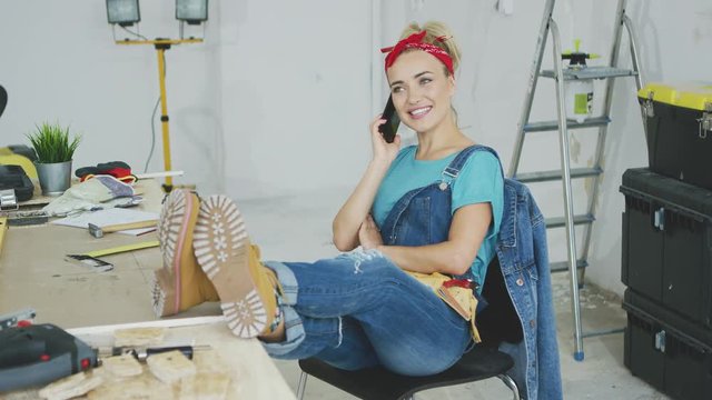 Cheerful young blond woman in jeans overalls and tool belt sitting comfortably with legs on carpenter desk talking on mobile phone with stretched out hand and looking up smiling.