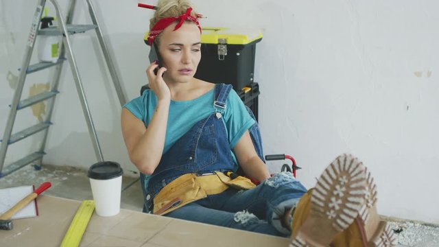 Young blond woman in jeans overalls sitting with legs on top of wooden desk with tools and talking on mobile phone anxiously looking away with white plastered wall and stepladder on background .