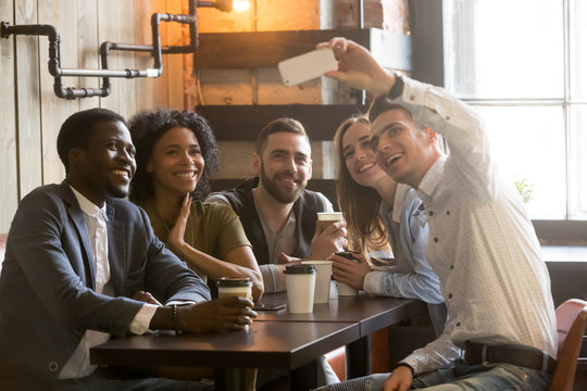 Smiling Caucasian man taking self-portrait on smartphone with multiracial friends sitting in coffee shop, diverse people shooting, making selfie on phone camera, chilling having fun out in cafe