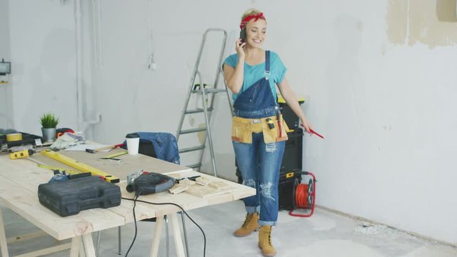 Happy smiling young blond woman in jeans overalls standing at carpenter workbench at unpainted wall with stepladder and instruments talking on mobile phone and looking away .