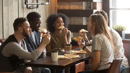 Foto op Aluminium Glimlachende multiraciale vrienden die pizza eten en koffie drinken, lachen en plezier hebben in restaurant, diverse millenniumcollega& 39 s genieten van lunch tijdens werkpauze zittend aan salontafel in loft café © fizkes