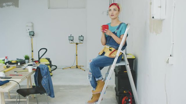 Attractive young blond woman in jeans overalls, tool belt and red headband sitting relaxed on stepladder holding red mug with drink and looking away dreamily at unpainted wall and workbench