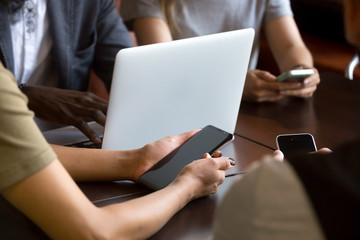Close up of multiracial people using gadgets sitting at table, working on laptops, smartphones, reading news or checking email, diverse technology obsessed workers addicted to devices during meeting.