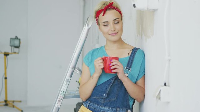 Beautiful blond woman in jeans overalls and red headband standing relaxed with mug of hot drink leaning on shoulder against white unpainted wall and looking at camera smiling.