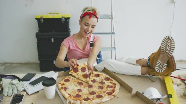 Beautiful young blond smiling female in pink shirt and white overalls sitting with legs on workbench and taking piece of delicious pizza 