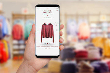 Female hand holding smartphone and shopping online, blurred clothing store in background. Online...