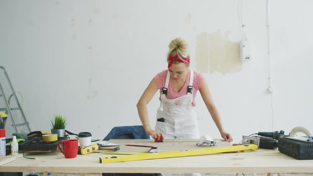 Young blond woman in pink shirt and white overalls with tape measure bending over wooden workbench with tools checking length of plank on background of white unpainted wall .