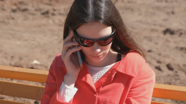 Young brunette girl in sunglasses waiting for a call on the phone sitting in the Park on the bench.