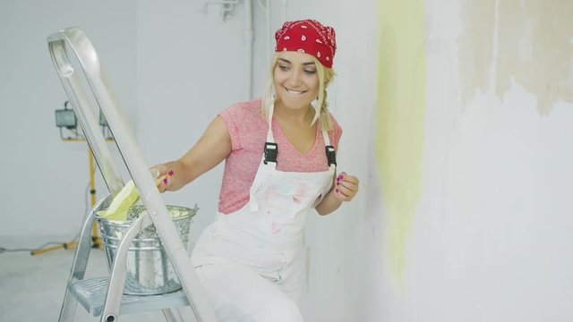 Beautiful young blond woman in white overalls and red bandana dipping brush into metal bucket with pastel yellow wall paint placed on top of stepladder 