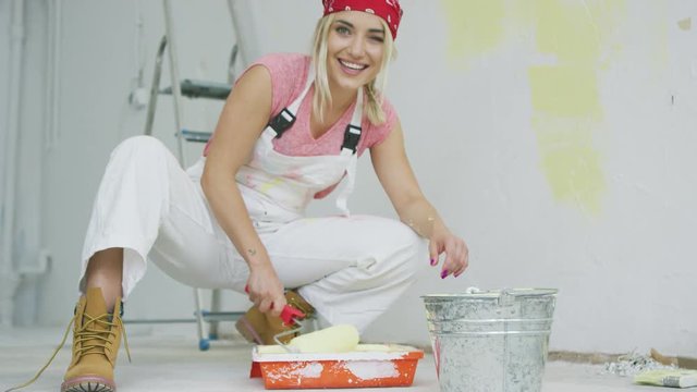 Crop view of happy young attractive woman in white overalls and red bandana sitting on hunkers at bucket and dipping roller in tray with pastel yellow wall paint looking at camera and smiling.